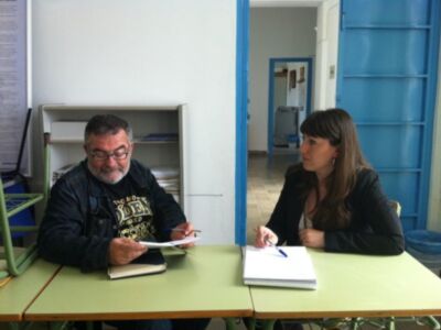 Un hombre y una mujer sentados en una mesa de oficina, ambos escribiendo en cuadernos. El ambiente es profesional y ordenado, con estanterías y un refrigerador en el fondo.