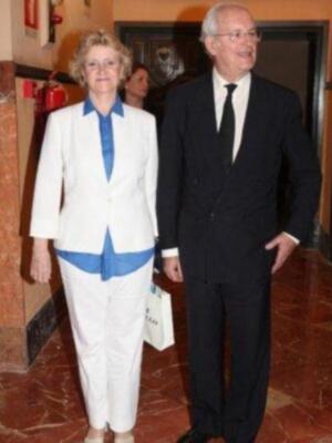 Una mujer con un traje blanco y azul, junto a una persona en traje negro, posando frente a una puerta de madera.