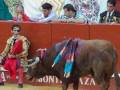 Un torero con un traje tradicional de color rojo y amarillo se encuentra sentado en una silla, mientras un toro con cuernos y adornos de color azul y rojo lo observa. En el fondo, varios espectadores observan la escena en un ambiente festivo con banderas y decoraciones.