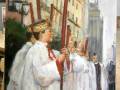 Pintura que muestra a una figura vestida con un manto blanco y tocado, seguida por otros personajes en traje religioso, todos con velas rojas. El fondo muestra una escena urbana con edificios y árboles.