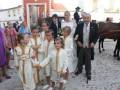 Una boda tradicional en un pueblo con niños vestidos de blanco y dorado, adultos en trajes formales, y una pareja casada a caballo.
