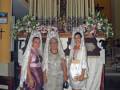Tres mujeres vestidas de blanco posan frente a un altar adornado con velas y flores, probablemente durante una celebración religiosa.
