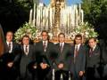 Un grupo de hombres en trajes formales se encuentra junto a una estatua religiosa, rodeados por flores y velas. El cielo es claro y azul.