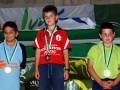 Tres niños posan con medallas en un podio, vestidos con camisetas de diferentes equipos. Fondo con banderas y logotipos deportivos.