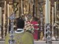 Un sacerdote en vestimenta religiosa, frente a una imagen de la Virgen María, con flores y velas en el altar.