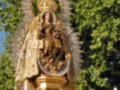 Una estatua de la Virgen María en un altar, rodeada de flores y decoraciones doradas. La escena parece ser parte de una celebración religiosa o festividad, con árboles y cielo azul en el fondo.