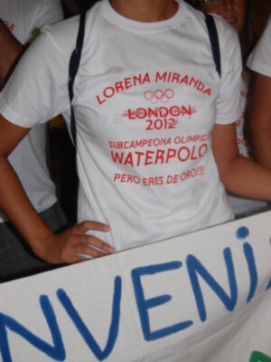 A una persona con un camiseta blanca que dice "LORENA MIRANDA LONDON 2012 SUBCAMPEONA OLIMPICA WATERPOLO PERO ERES DE ORO!!" y detrás hay una pancarta con letras azules.
