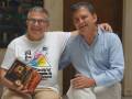 Dos hombres sonrientes, uno sosteniendo un libro con una portada de color. El fondo muestra una habitación interior con muebles y decoraciones.