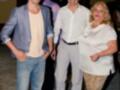 Tres personas posando juntas en una fiesta nocturna, dos hombres y una mujer. El hombre de la izquierda lleva un blazer azul claro, el hombre del medio está con una camisa blanca y pantalones grises, mientras que la mujer de la derecha lleva una camisa blanca y pantalones con estampado animal.