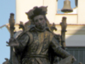 La estatua de bronce muestra a un personaje histórico con una corona y vestimenta medieval, sosteniendo un libro abierto en una mano. La imagen se centra en la estatua, con el fondo incluyendo un edificio con una campana visible.