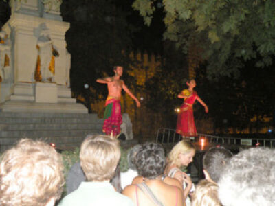 Un hombre y una mujer están bailando en un escenario iluminado por velas, mientras una multitud observa atentamente. La escena se desarrolla en la noche frente a un monumento histórico, con árboles y una estructura de piedra en el fondo.