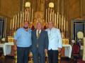 Tres hombres posan en una iglesia con un altar y velas.