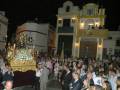 Una procesión religiosa nocturna en una ciudad con iglesia de estilo colonial, gente vestida formalmente y flores en el altar.