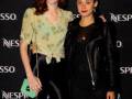 Dos mujeres posan en un evento Nespresso, con una de ellas usando un top verde floral y pantalones negros, mientras la otra lleva una chaqueta de cuero negra y un vestido negro.