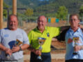 Tres hombres celebran un triunfo en una competición deportiva, posando con trofeos y sonriendo. El ambiente es al aire libre, con un paisaje de montañas en el fondo y una valla metálica alrededor del campo.