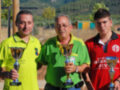 Tres personas posan con trofeos en un entorno deportivo, con árboles y montañas al fondo.