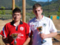 Dos jóvenes posan con trofeos, uno en rojo y otro en blanco. El fondo muestra un campo de juego con árboles y montañas al fondo, bajo un cielo azul.