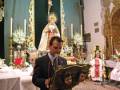 Un hombre en traje sostiene un libro de oraciones frente a una imagen religiosa en un altar. La decoración incluye flores y otros elementos de arte religioso.