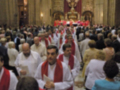 Una multitud de personas en una iglesia, algunos con vestidos blancos y otros con colores. La imagen muestra a varios sacerdotes en el centro, rodeados de fieles. En la parte superior se puede ver un altar con una imagen central y una cruz en el fondo.