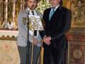 Dos hombres en trajes formales, uno con corbata y el otro con un cuello alto, posan frente a una araña de cristal. El fondo muestra detalles de la decoración interior, incluyendo un altar con flores y una estatua.