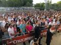 Un grupo numeroso de personas en un estadio, con una gran cantidad de espectadores sentados y otros caminando. La imagen muestra a varios individuos, incluyendo un hombre en una camiseta azul y otro en uniforme militar. El fondo muestra árboles y una estructura de iluminación. La imagen está firmada con "Cruzcampo" en el lado izquierdo de la barandilla.