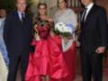 Una mujer vestida de rojo con un vestido de gala y una corona, posa junto a dos hombres en trajes formales. La escena parece ser un evento formal, posiblemente una boda o recepción de gala.