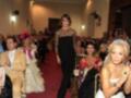 Una mujer camina por una pasarela en un evento formal, mientras un público aplaude y una mujer con un vestido blanco y un diamante en la mano la observa.