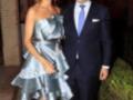 Una pareja posa en un evento formal. La mujer lleva un vestido plateado con detalles de encaje y el hombre está formalmente vestido con una chaqueta azul claro. La escena parece ser al aire libre, posiblemente en una entrada de edificio histórico.