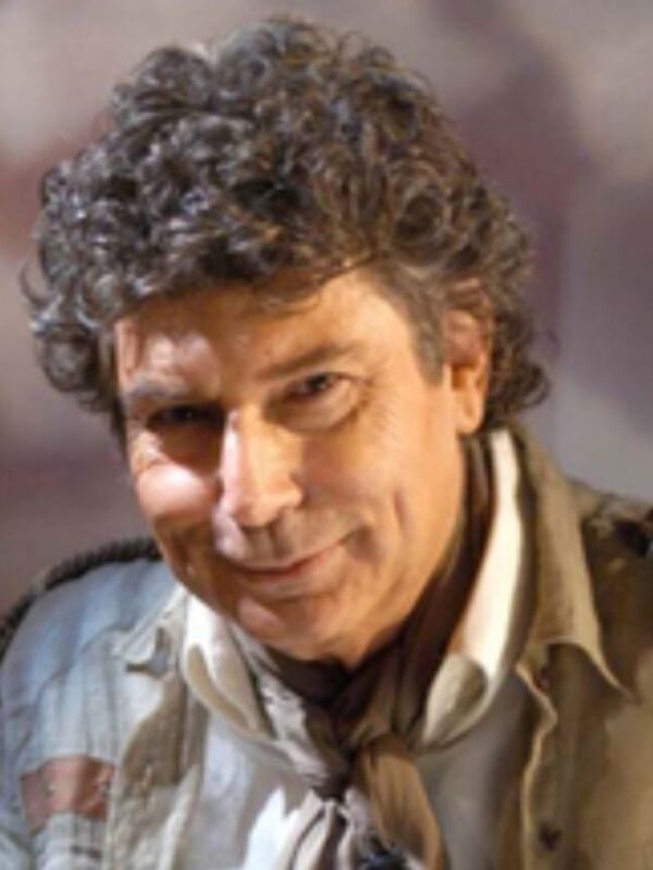 Un hombre con cabello oscuro y rizado, vistiendo una chaqueta beige y un pañuelo en el cuello.