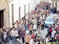 Una multitud de personas camina en fila por una calle estrecha, con edificios blancos a ambos lados. Algunas personas llevan bolsas y carros de bebé, mientras otros parecen estar esperando algo. La escena parece ser una procesión o reunión importante, con la gente apretada y animada.