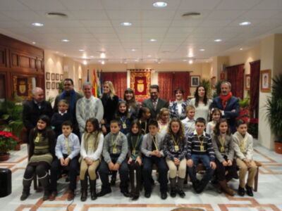 Una foto de grupo en un salón con decoración formal. La imagen muestra a varias personas sentadas y posando para la foto, incluyendo adultos y niños. La decoración del salón incluye una bandera, un escudo y plantas en macetas. La iluminación es suave, con luces de techo y ventanas que permiten la entrada de luz natural.