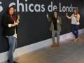 Tres personas celebran en una presentación de "Las Chicas de Londres". Una sostiene un trofeo, otra muestra una victoria con la mano izquierda y la tercera sonríe ampliamente. El fondo es una gran pared negra con letras blancas y un logo en la parte superior derecha.