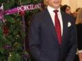 Elegante hombre en traje y corbata roja posando frente a un árbol de Navidad decorado con luces y adornos rojos y violetas.