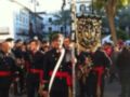 Imagen de un desfile militar con soldados en uniforme negro y rojo, sosteniendo banderas. El fondo muestra edificios europeos con balcones y palmeras, bajo un cielo despejado.