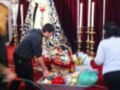 Una imagen festiva con una figura de la Virgen en un altar dorado, rodeada por velas y candelabros. Una persona pone una carretilla con regalos en el suelo rojo, mientras otra observa.