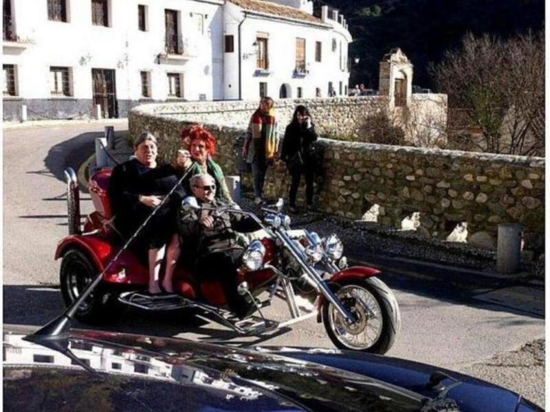 Una familia disfruta de una aventura en un triciclo motorizado, rodeados por un ambiente rural con una casa blanca y un muro de piedra en el fondo.