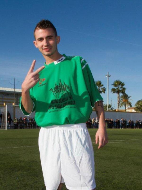 Un joven con una camiseta verde y pantalones blancos posa en un campo de fútbol, con palmeras y una estructura blanca al fondo.