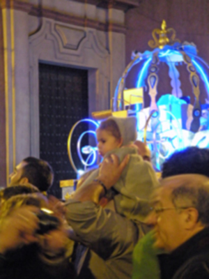 Una imagen captura una celebración festiva con un niño en brazos, disfrutando de la fiesta. La escena está ambientada en una calle con edificios antiguos y un círculo azul brillante que parece ser parte de una decoración navideña o festiva. La iluminación y el ambiente sugieren un evento especial en una ciudad histórica o turística.
