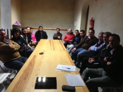 Una reunión en un salón con una mesa de madera, personas sentadas alrededor, algunos con portátiles y documentos en la mesa.