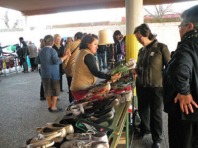 Una persona está comprando zapatos en un puesto de mercado al aire libre. El puesto tiene una variedad de calzado visible y está rodeado por otros compradores y vendedores. La escena se encuentra bajo una estructura de toldo, con gente caminando y conversando en el fondo.