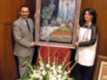 Dos personas sostienen una pintura de la Virgen de Guadalupe, rodeadas por flores blancas y un marco dorado. La escena transmite una sensación de devoción y celebración religiosa.
