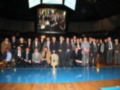Una reunión formal en un estadio de baloncesto, con una gran pantalla en el fondo mostrando a varios hombres. La mayoría están vestidos formalmente, algunos con corbatas y otros con chalecos. Algunos hombres llevan medallas o insignias, indicando posiblemente un reconocimiento o premio. La escena transmite una sensación de orgullo y celebración entre los participantes.
