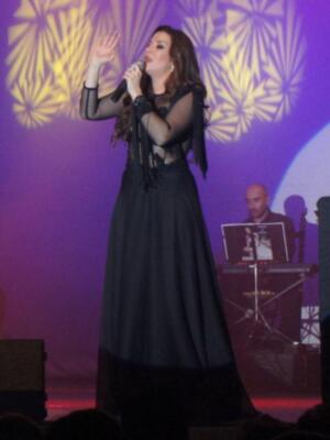 Una mujer cantante en un escenario con una vestimenta negra y larga, sosteniendo un micrófono. En la pantalla de fondo se puede ver a un músico tocando teclados.