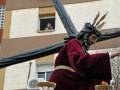 Un hombre vestido con un manto rojo y una corona de espinas, sostiene un poste largo en la procesión. Detrás se ve una fachada de edificio con ventanas y un balcón.