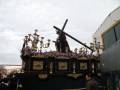 Imagen de una procesión religiosa con un paso que porta una figura en la cruz, rodeada por flores y adornos dorados. El cielo es nublado y hay una estructura industrial en el fondo.