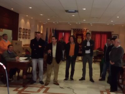 Una reunión formal en un salón con una mesa y sillas, varios hombres de negocios vestidos formalmente, algunos con chaquetas negras y otros con camisas coloridas. La decoración incluye banderas y un escudo en el fondo, iluminación de techo y cortinas rojas.