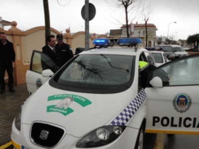Un policía local de Chipiona dirige su coche blanco con logotipo verde y azul, mientras otros agentes observan. Fondo urbano con edificios y coches en el camino.
