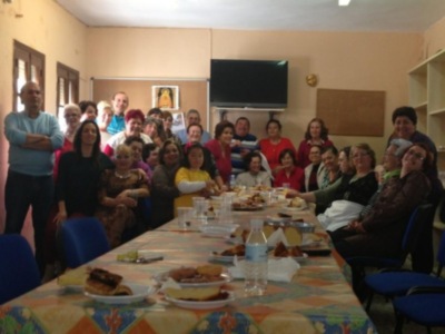 Grupo de personas sentadas en una sala comedor, con mesa llena de alimentos y sillas alrededor. En la pared hay un televisor, una caja de madera y un cartel. La sala tiene un ambiente familiar y alegre.