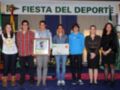 Fiesta del Deporte celebrada en un escenario con banderas y decoración. Un grupo de personas, entre ellos una persona sosteniendo un diploma con el título "Premio a la Excelencia Académica". La imagen muestra una celebración deportiva y académica.
