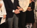 Una imagen de una gala o evento formal donde un hombre y una mujer están saludándose. El hombre lleva un traje oscuro con corbata, mientras que la mujer está vestida con una blusa de manga larga y pantalones negros. Ambos están sonriendo y parecen estar en un ambiente de celebración o reconocimiento.